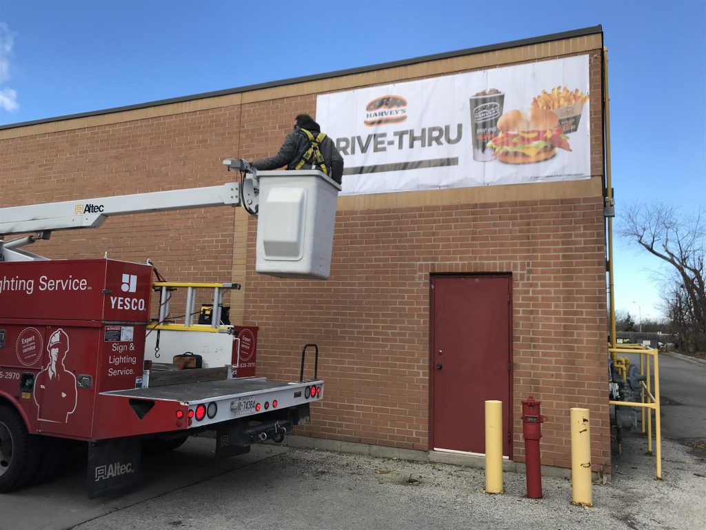 a photo of the drive thru sign on the side of Harvey's restaurant showing food. the Yesco technician is in the boom of the truck working on the sign.